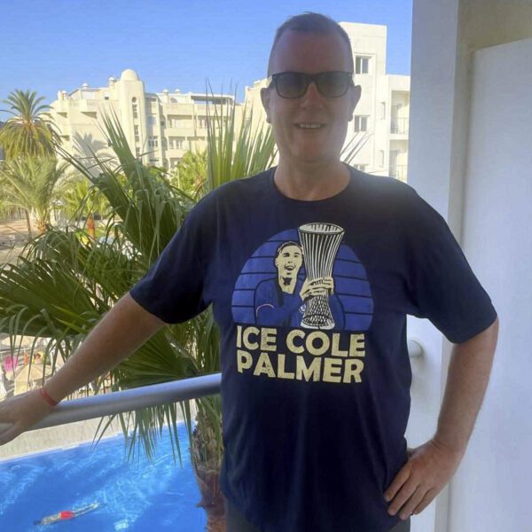 A smiling man wearing a navy tee of Ice Cole Palmer lifting the Conference League trophy for Chelsea.