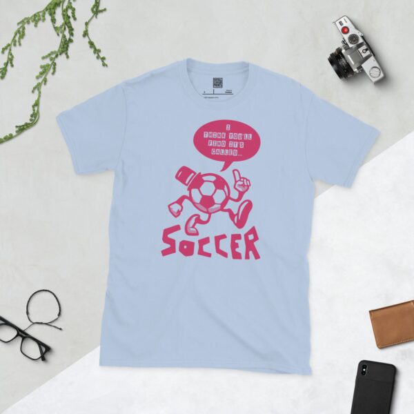 A baby blue T-shirt featuring a pink cartoon football character with a top hat, arms, and legs, pointing. A speech bubble above reads "I THINK YOU'LL FIND IT'S CALLED..." and "SOCCER" is written below the character.