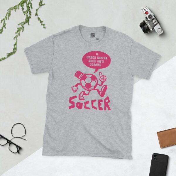 A sports grey T-shirt featuring a pink cartoon football character with a top hat, arms, and legs, pointing. A speech bubble above reads "I THINK YOU'LL FIND IT'S CALLED..." and "SOCCER" is written below the character.