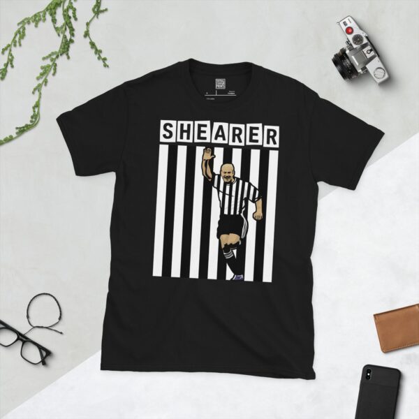 Black: A bold black T-shirt featuring an illustration of Alan Shearer in his iconic Newcastle United black and white striped kit, arm raised in celebration. His name "SHEARER" is prominently displayed in white blocks above him against a striped background.