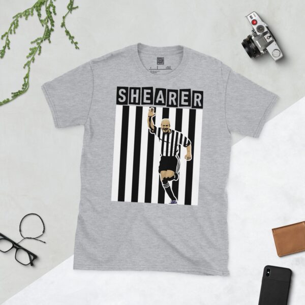 A versatile sports grey T-shirt featuring a striking illustration of Alan Shearer, the revered Newcastle United striker, in his traditional black and white kit with his arm raised in triumph. His surname "SHEARER" is positioned above the graphic, completing the tribute.