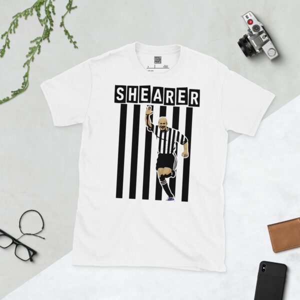 A crisp white T-shirt showcasing a dynamic graphic of Newcastle United legend Alan Shearer in his famous black and white striped shirt, mid-celebration with his arm raised. The surname "SHEARER" is clearly visible in block letters above the design.