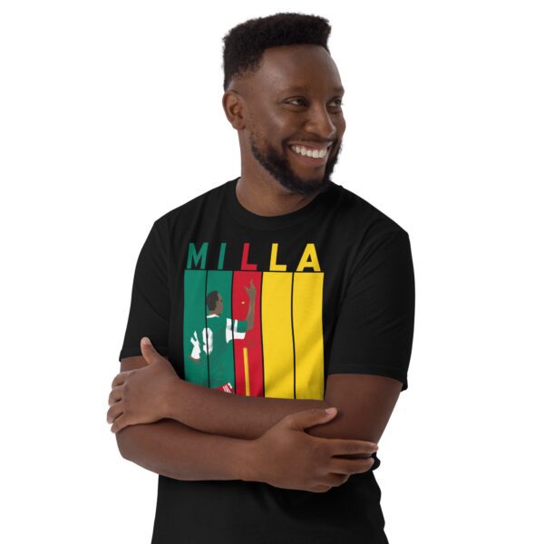 A smiling black man wearing a black Roger Milla T-shirt featuring a bold striped design in Cameroon flag colours, with "MILLA" above five pillars and a graphic of Roger Milla doing his iconic corner flag dance from the 1990 World Cup.