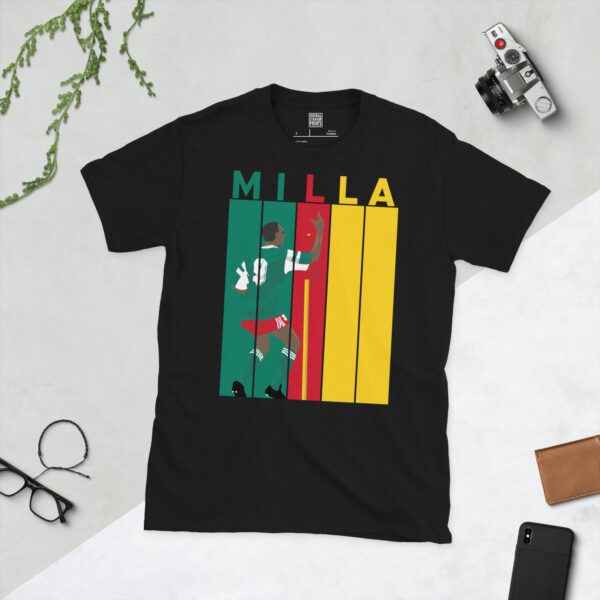 A black Roger Milla T-shirt featuring a bold striped design in Cameroon flag colours, with "MILLA" above five pillars and a graphic of Roger Milla doing his iconic corner flag dance from the 1990 World Cup.