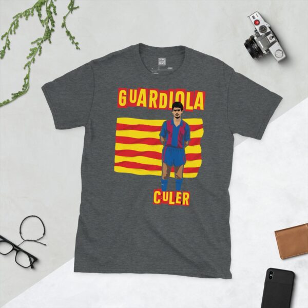A subtle dark heather T-shirt displaying a distinctive graphic of former Barcelona star Pep Guardiola as a young player. His portrait is captured against a retro Catalan flag backdrop, with the beloved term "CULER" directly beneath the illustration.