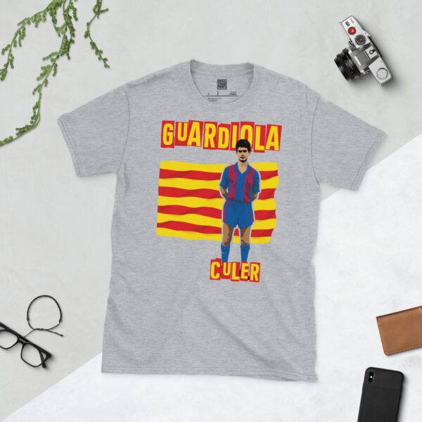 A versatile sports grey T-shirt showcasing a dynamic illustration of a young Pep Guardiola, the beloved Barcelona midfielder. The design includes his full figure over a vibrant red and yellow striped flag, with "CULER" clearly visible underneath.