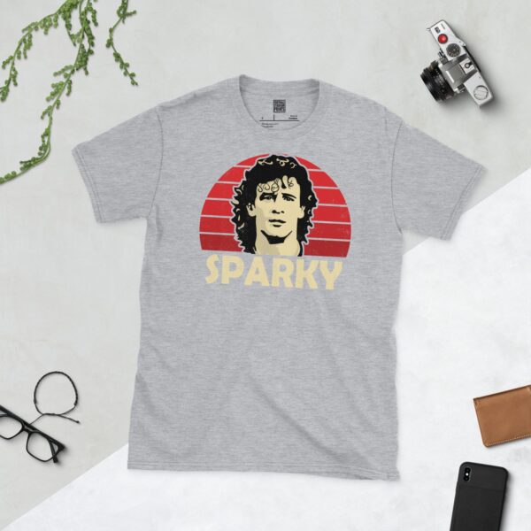 A versatile sports grey T-shirt showcasing a dynamic illustration of Mark Hughes, known as 'Sparky'. The design includes his portrait over a bold red radiating pattern, with "SPARKY" clearly visible underneath, celebrating the iconic player.