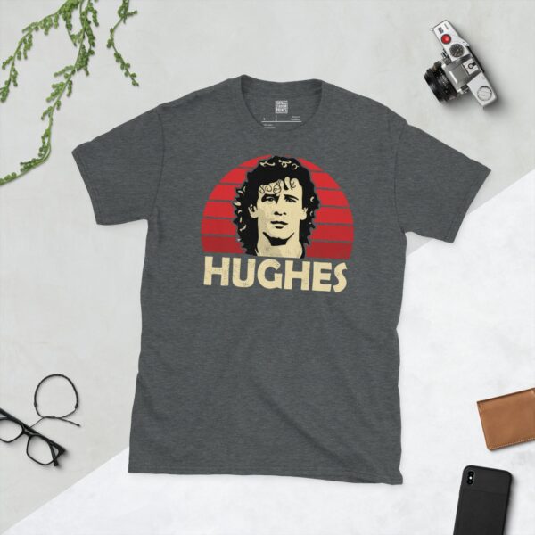 A subtle dark heather T-shirt displaying a distinctive graphic of former football star Mark Hughes. His portrait is captured against a retro red sunburst effect, with his surname "HUGHES" directly beneath the illustration.