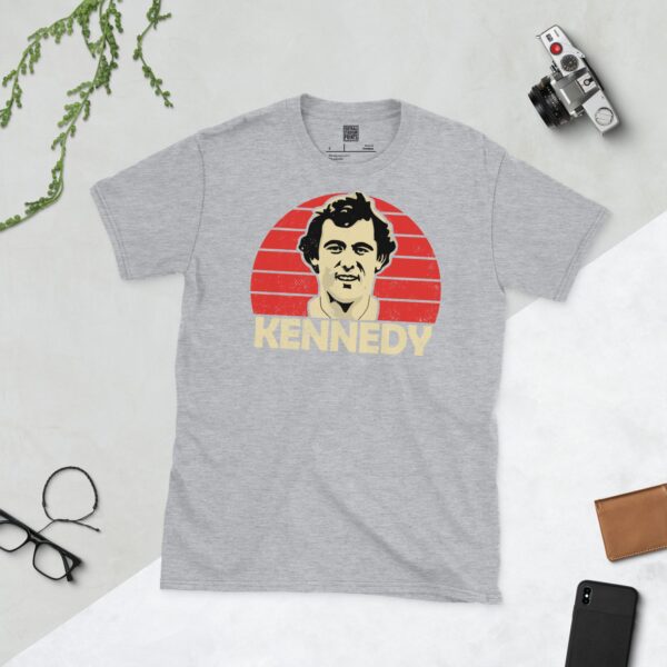A versatile sports grey T-shirt showcasing a dynamic illustration of Ray Kennedy, the memorable Liverpool midfielder. The design includes his portrait over a bold red radiating pattern, with "KENNEDY" clearly visible underneath.