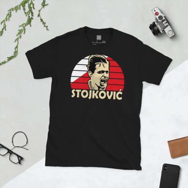 A bold black T-shirt emblazoned with a striking portrait of Red Star Belgrade icon Dragan Stojković. The graphic depicts his face against a backdrop of red and white sunburst lines, and his surname "STOJKOVIĆ" is a central feature below the image.