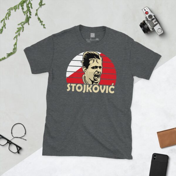 A subtle dark heather T-shirt displaying a distinctive graphic of former Red Star Belgrade maestro Dragan Stojković. His portrait is captured against a retro red and white sunburst effect, with his surname "STOJKOVIĆ" directly beneath the illustration.