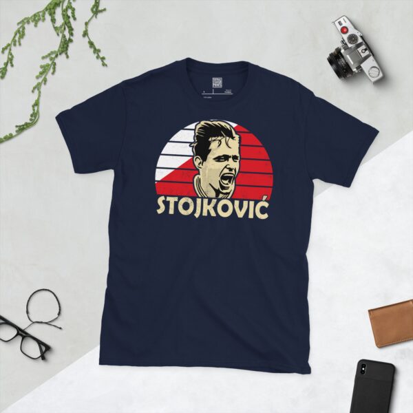 A deep navy blue T-shirt featuring a compelling graphic of Dragan Stojković, the legendary Red Star Belgrade forward, with his image set against a vivid red and white striped design. His iconic surname "STOJKOVIĆ" is clearly visible, paying tribute to his time at the club.