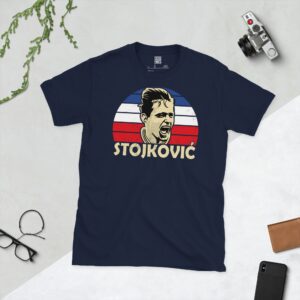 A deep navy blue T-shirt featuring a compelling graphic of Dragan StojkoviΔ, the legendary Yugoslavian forward, with his image set against a vivid blue, white, and red striped design. His iconic surname "STOJKOVIΔ" is clearly visible, paying tribute to his time with the national team.