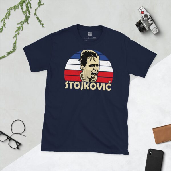 A deep navy blue T-shirt featuring a compelling graphic of Dragan Stojković, the legendary Yugoslavian forward, with his image set against a vivid blue, white, and red striped design. His iconic surname "STOJKOVIĆ" is clearly visible, paying tribute to his time with the national team.