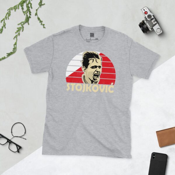 A versatile sports grey T-shirt showcasing a dynamic illustration of Dragan Stojković, the beloved Red Star Belgrade midfielder. The design includes his portrait over a bold red and white radiating pattern, with "STOJKOVIĆ" clearly visible underneath.