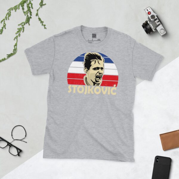 A versatile sports grey T-shirt showcasing a dynamic illustration of Dragan Stojković, the beloved Yugoslavian midfielder. The design includes his portrait over a bold blue, white, and red radiating pattern, with "STOJKOVIĆ" clearly visible underneath.