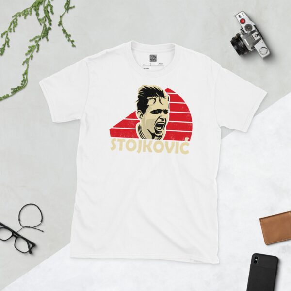 A clean white T-shirt featuring a stylized graphic of football legend Dragan Stojković, set against a circular red and white striped background inspired by the Red Star Belgrade badge. His surname "STOJKOVIĆ" is prominently displayed below the portrait.