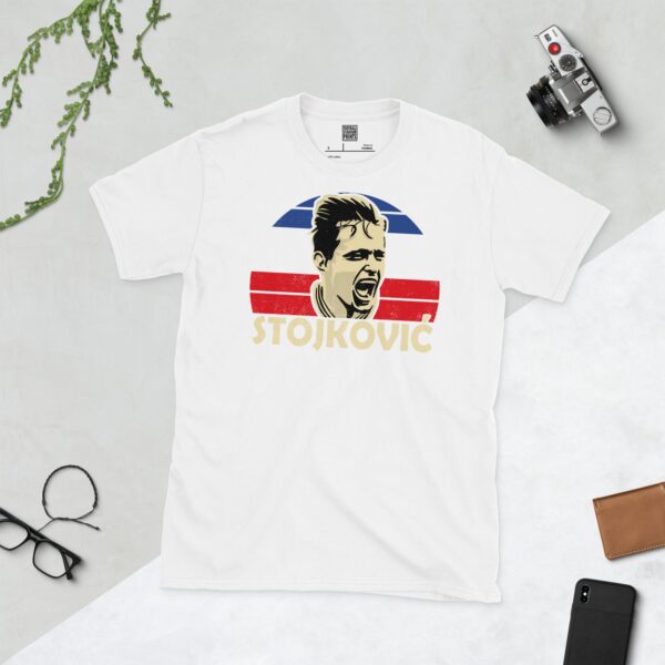 A clean white T-shirt featuring a stylized graphic of football legend Dragan Stojković, set against a circular background of blue, white, and red stripes inspired by the former Yugoslavia flag. His surname "STOJKOVIĆ" is prominently displayed below the portrait.