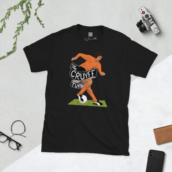 A bold black T-shirt emblazoned with a striking illustration of Johan Cruyff performing his iconic 'Cruyff Turn'. The graphic features his full figure in a bright orange kit and is a tribute to his legacy, with "THE CRUYFF TURN" prominently featured.