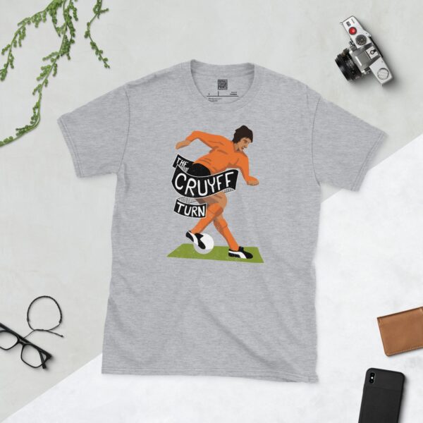 A versatile sports grey T-shirt showcasing a dynamic illustration of football legend Johan Cruyff, mid-move during his iconic 'Cruyff Turn'. The graphic highlights the famous technique with the text "THE CRUYFF TURN" in a retro font.