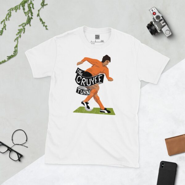 A clean white T-shirt featuring a stylized illustration of Johan Cruyff in a retro orange kit, performing his famous 'Cruyff Turn'. The phrase "THE CRUYFF TURN" is displayed in bold text, celebrating his legendary status.