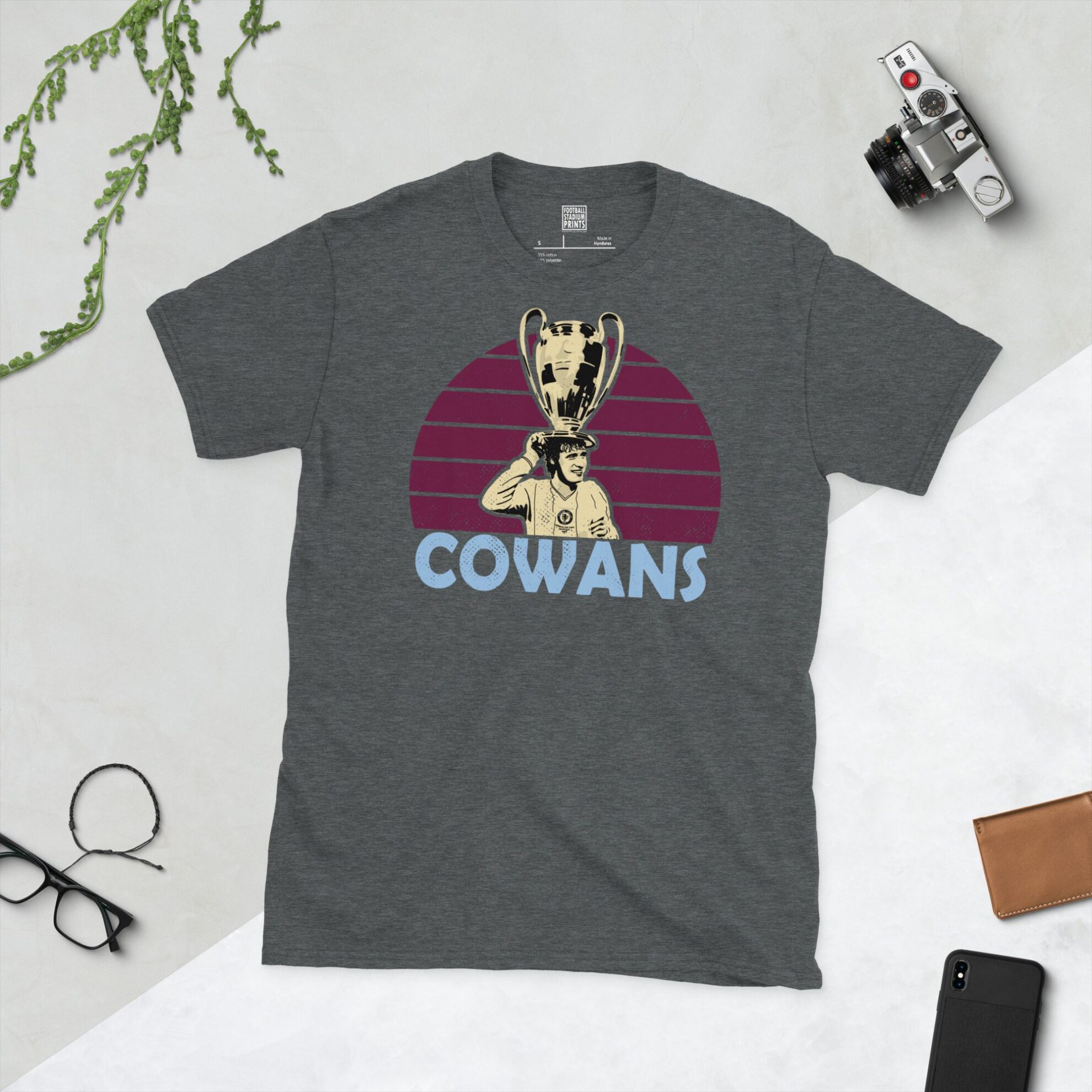 A subtle dark heather T-shirt displaying a distinctive graphic of former Aston Villa maestro Gordon Cowans. His portrait shows him lifting the European Cup, captured against a retro claret sunburst effect, with his surname "COWANS" directly beneath the illustration.