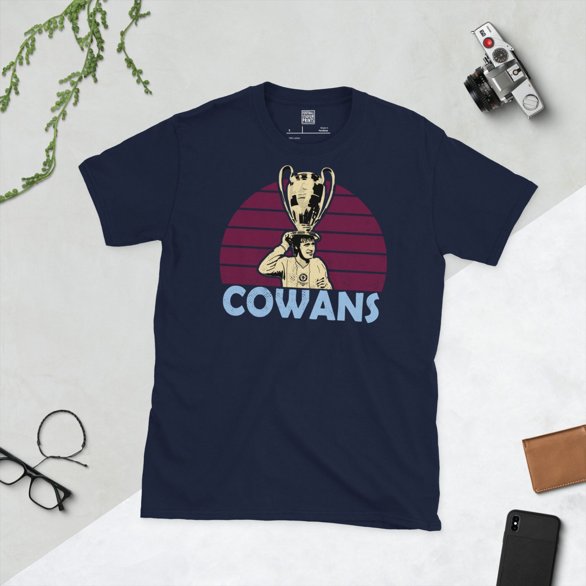 A deep navy blue T-shirt featuring a compelling graphic of Gordon Cowans, the legendary Aston Villa midfielder, with his image lifting the European Cup set against a vivid claret sunburst design. His iconic surname "COWANS" is clearly visible, paying tribute to his time at the club.