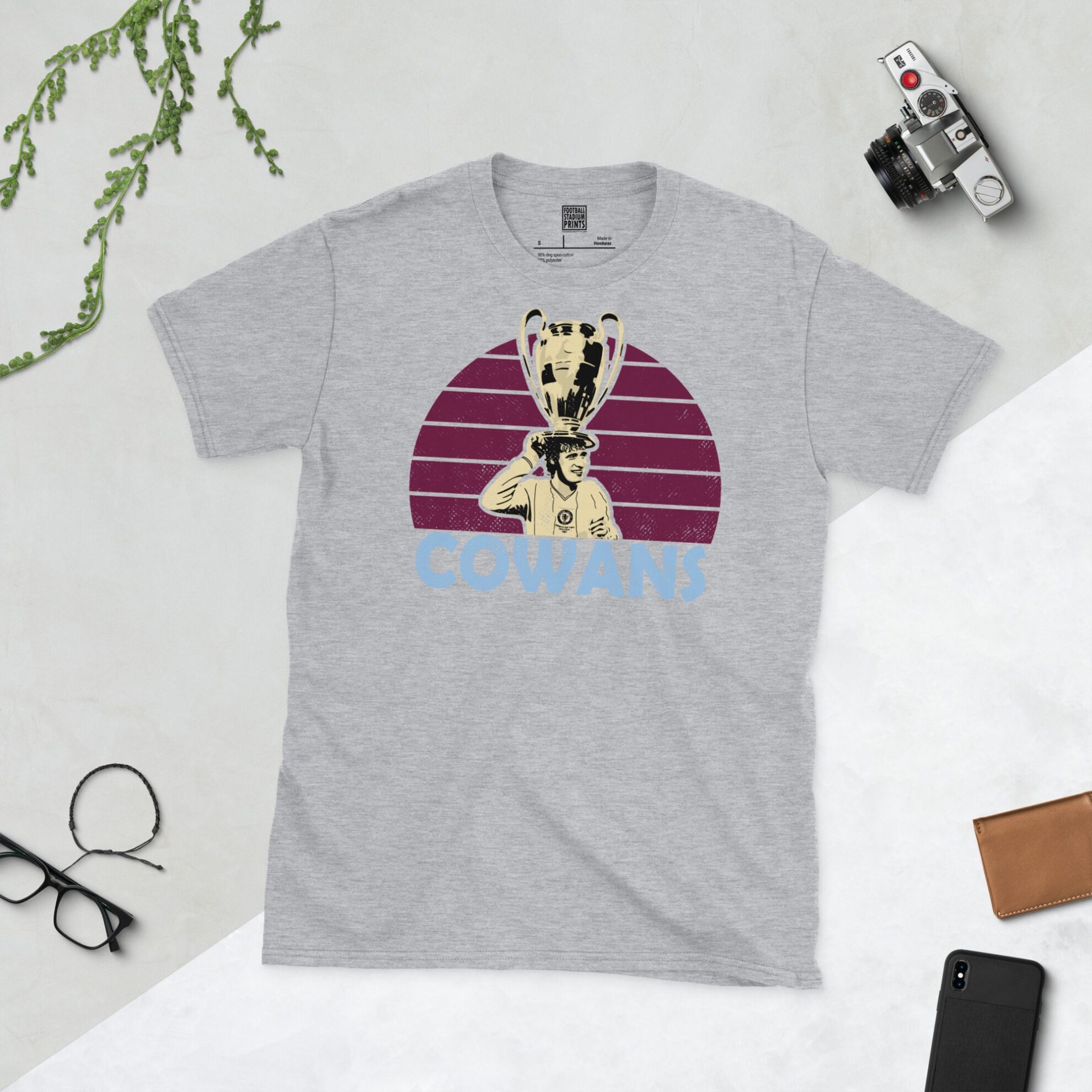 A versatile sports grey T-shirt showcasing a dynamic illustration of Gordon Cowans, the beloved Aston Villa midfielder and European Cup winner. The design includes his portrait with the trophy over a bold claret radiating pattern, with "COWANS" clearly visible underneath.