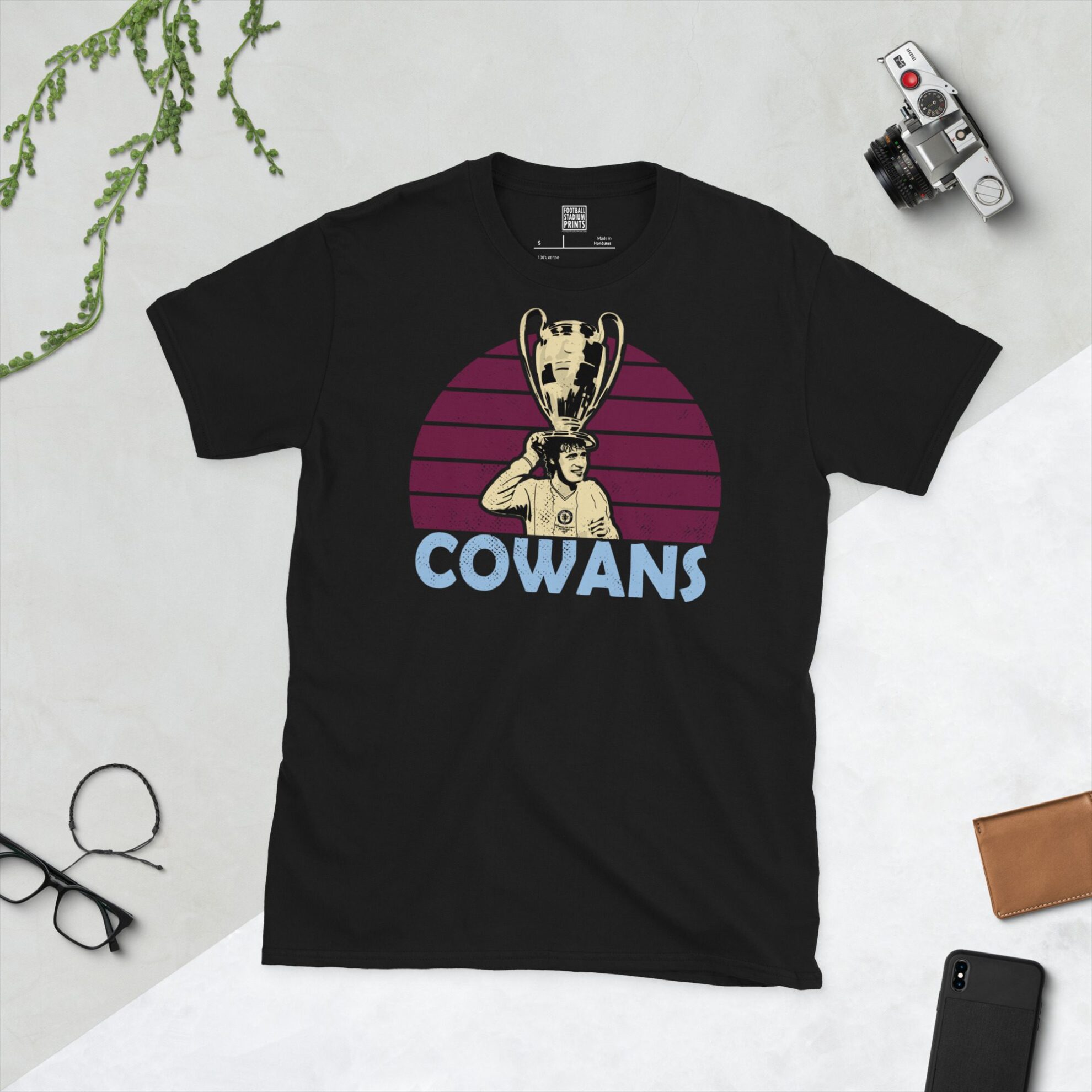 A bold black T-shirt emblazoned with a striking portrait of Aston Villa icon Gordon Cowans. The graphic depicts him lifting the European Cup against a backdrop of claret sunburst lines, and his surname "COWANS" is a central feature below the image.