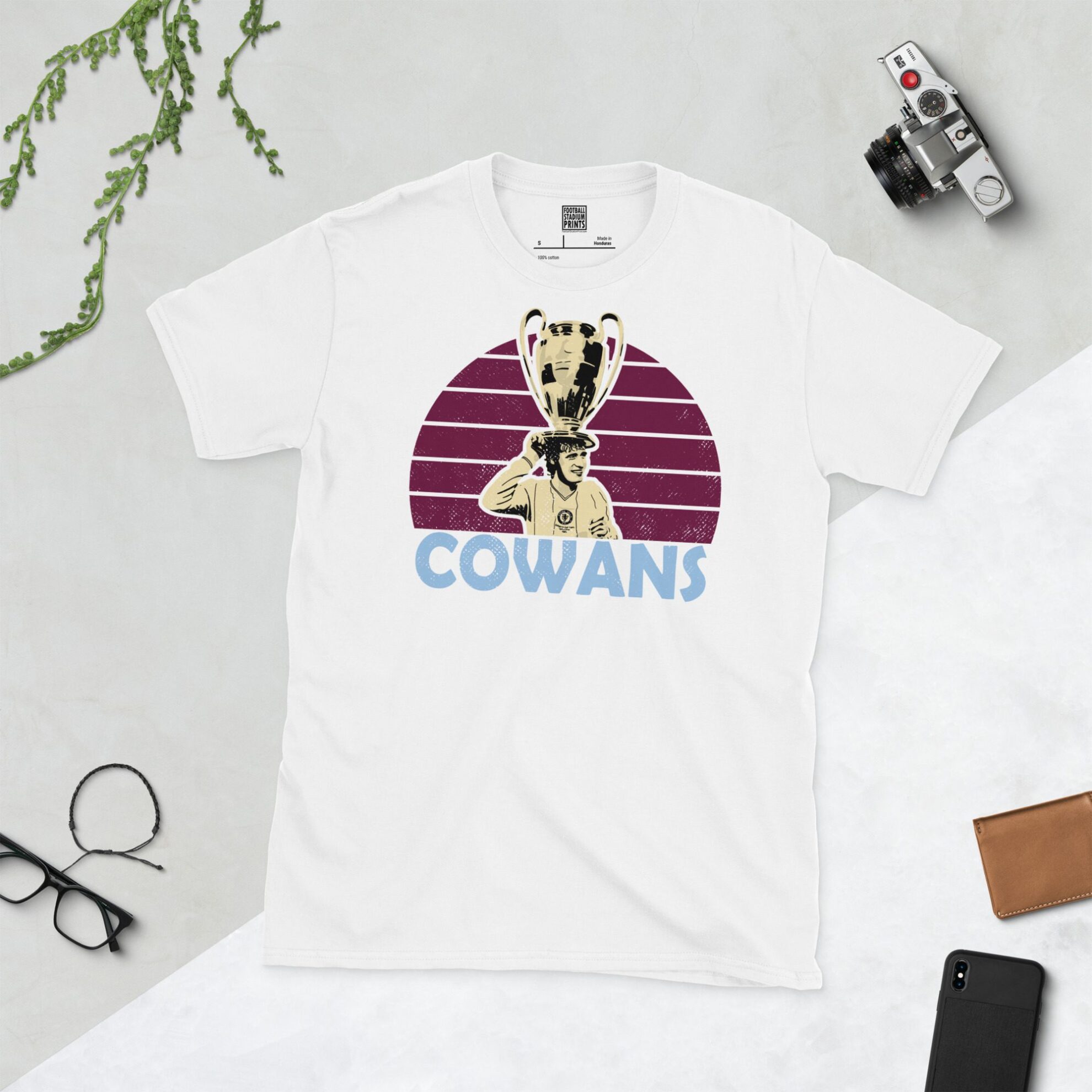 A clean white T-shirt featuring a stylised graphic of Aston Villa legend Gordon Cowans, holding the European Cup trophy, set against a vibrant sunburst background in a claret colour. His surname "COWANS" is prominently displayed below the portrait.