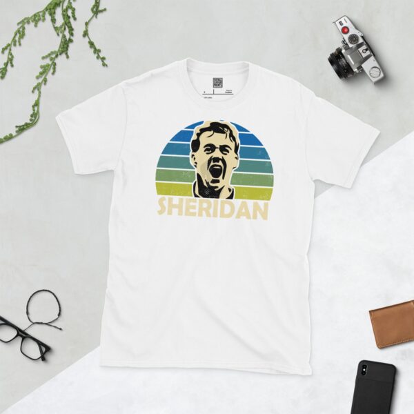 A clean white T-shirt featuring a stylised graphic of Leeds United legend John Sheridan, set against a vibrant sunburst background that fades from blue to yellow. His surname "SHERIDAN" is prominently displayed below the portrait.