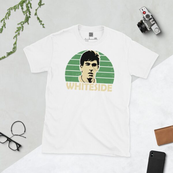 A clean white T-shirt featuring a stylised graphic of Northern Ireland legend Norman Whiteside's face, set against a vibrant sunburst background in green. His surname "WHITESIDE" is prominently displayed below the portrait.
