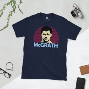 A deep navy blue T-shirt featuring a compelling graphic of Paul McGrath, the legendary Aston Villa defender, with his image set against a vivid claret sunburst design. His iconic surname "MCGRATH" is clearly visible, paying tribute to his time at the club.