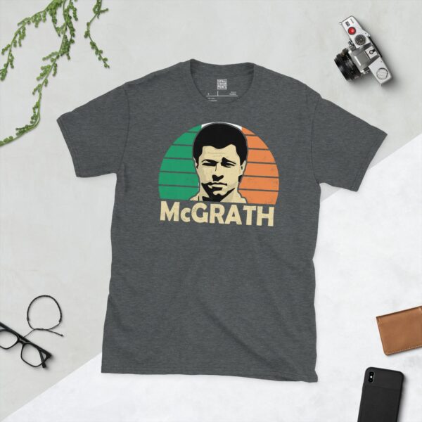 A subtle dark heather T-shirt displaying a distinctive graphic of former Republic of Ireland maestro Paul McGrath. His portrait is captured against a retro green, white, and orange sunburst effect, with his surname "MCGRATH" directly beneath the illustration.