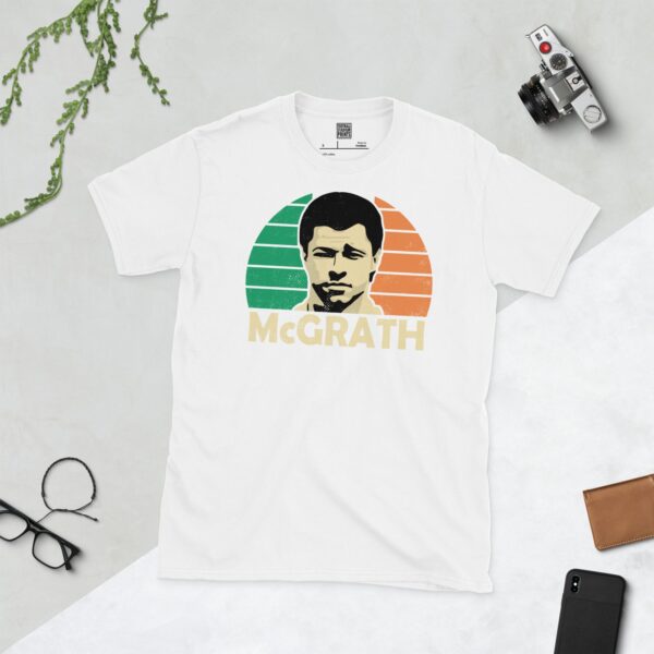 A clean white T-shirt featuring a stylised graphic of Republic of Ireland legend Paul McGrath's face, set against a vibrant sunburst background depicting the colours of the Irish flag (green, white, and orange). His surname "MCGRATH" is prominently displayed below the portrait.