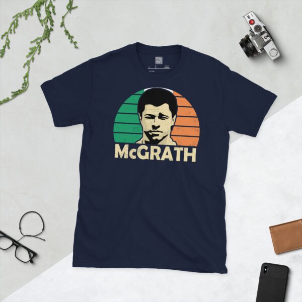 A deep navy blue T-shirt featuring a compelling graphic of Paul McGrath, the legendary Irish footballer, with his image set against a vivid green, white, and orange striped design. His iconic surname "MCGRATH" is clearly visible, paying tribute to his time with the national team.