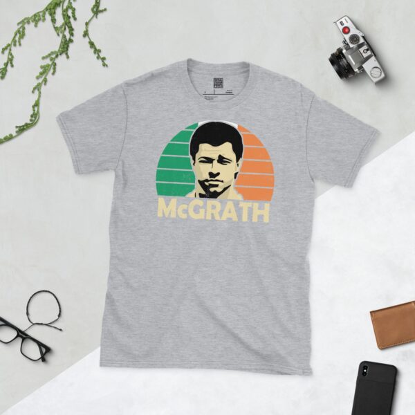 A versatile sports grey T-shirt showcasing a dynamic illustration of Paul McGrath, the beloved Irish national team defender. The design includes his portrait over a bold green, white, and orange radiating pattern, with "MCGRATH" clearly visible underneath.