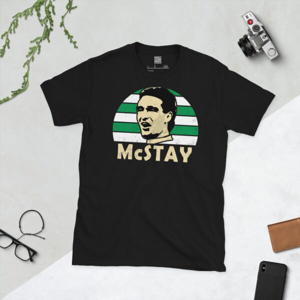 A bold black T-shirt emblazoned with a striking portrait of Celtic icon Paul McStay. The graphic depicts his face against a backdrop of green and white striped sunburst lines, and his surname "McSTAY" is a central feature below the image.