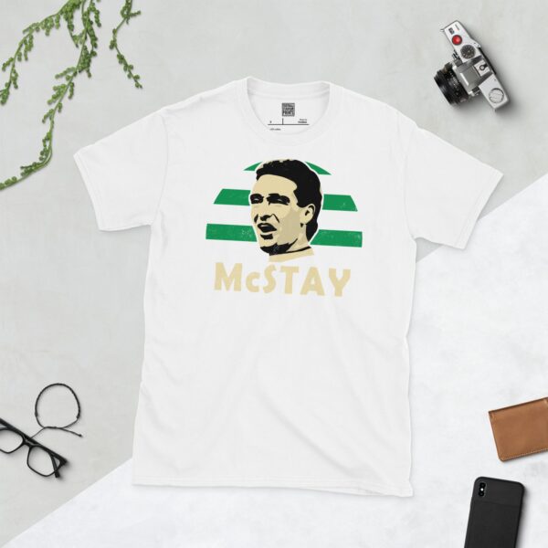 A clean white T-shirt featuring a stylised graphic of Celtic legend Paul McStay, set against a vibrant sunburst background in green and white stripes. His surname "McSTAY" is prominently displayed below the portrait.