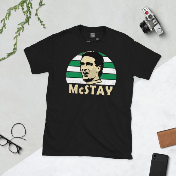 A bold black T-shirt emblazoned with a striking portrait of Celtic icon Paul McStay. The graphic depicts his face against a backdrop of green and white striped sunburst lines, and his surname "McSTAY" is a central feature below the image.