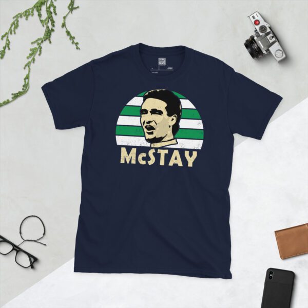 A deep navy blue T-shirt featuring a compelling graphic of Paul McStay, the legendary Celtic player, with his image set against a vivid green and white striped sunburst design. His iconic surname "McSTAY" is clearly visible, paying tribute to his time at the club.