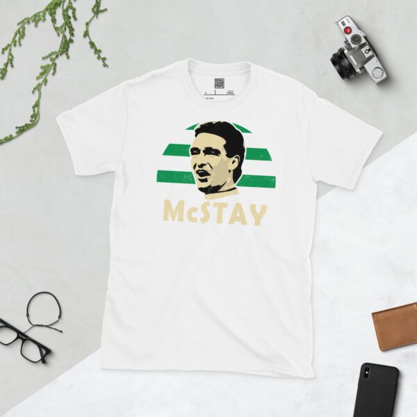 A clean white T-shirt featuring a stylised graphic of Celtic legend Paul McStay, set against a vibrant sunburst background in green and white stripes. His surname "McSTAY" is prominently displayed below the portrait.