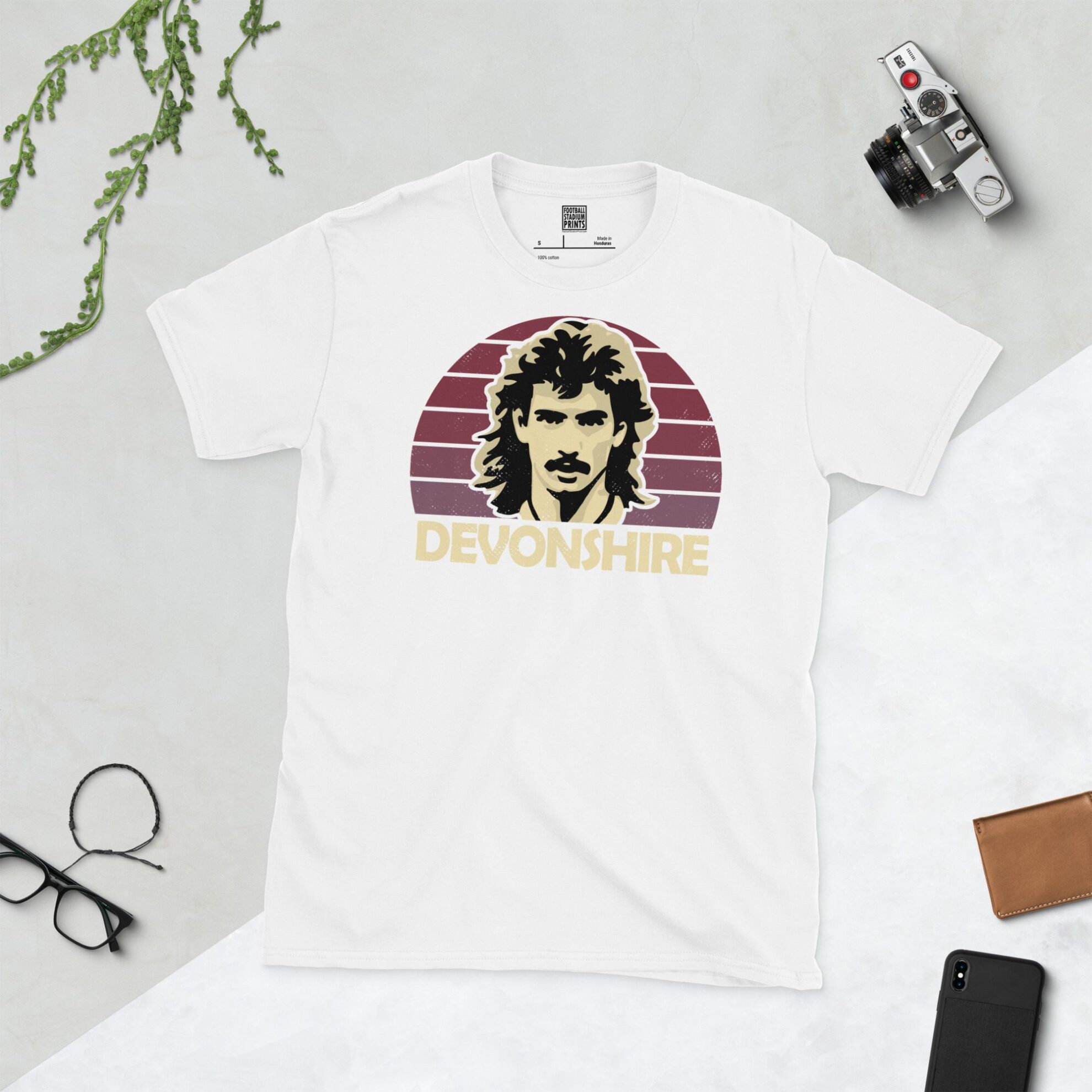 A white T-shirt with a bold portrait of football legend Alan Devonshire. The image is set against a claret sunburst background with the name "DEVONSHIRE" in light yellow underneath.