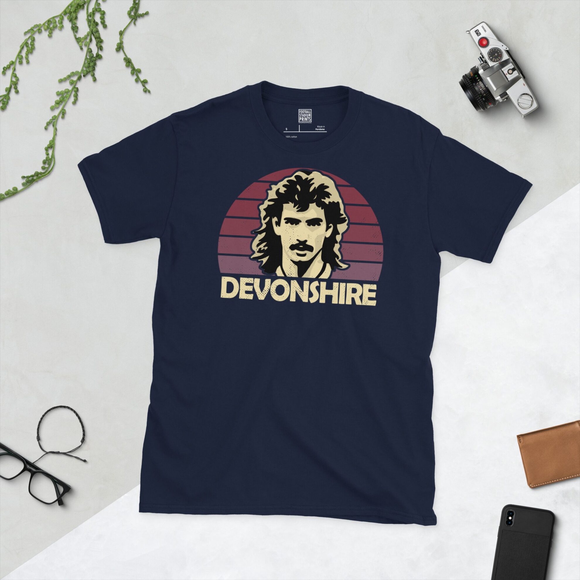 A navy T-shirt displaying an artistic illustration of Alan Devonshire, the former West Ham United midfielder. The graphic is backed by a claret sunburst and the name "DEVONSHIRE" appears underneath in light yellow.