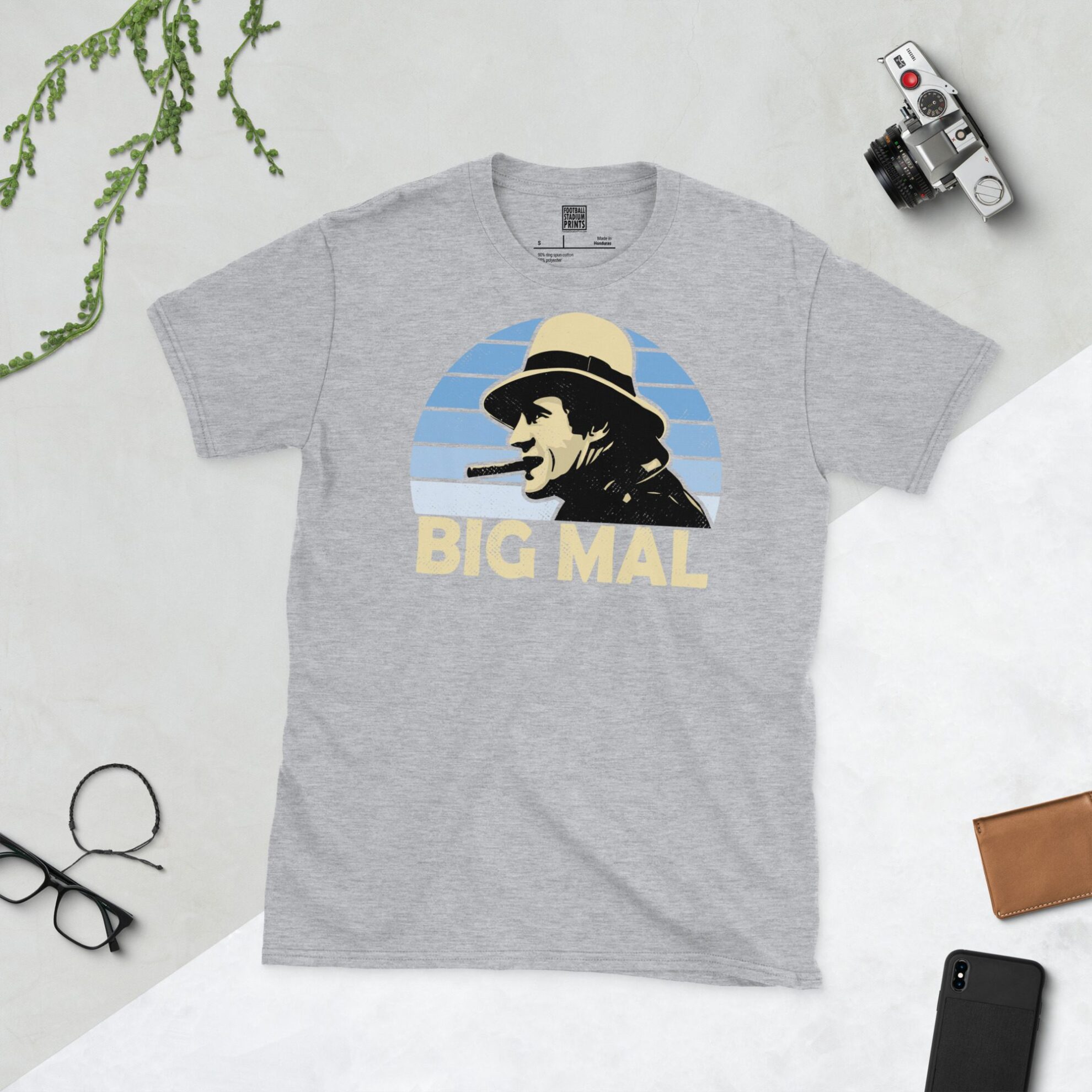 A sports grey T-shirt featuring a unique graphic of Man City's Malcolm Allison. The image is backed by a light blue faded sunburst and the text "BIG MAL" is printed in light yellow below the portrait.