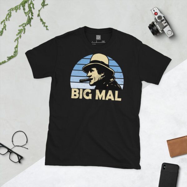 A black T-shirt featuring a stylised graphic of Manchester City legend Malcolm Allison, depicted with a fedora and cigar, against a light blue faded sunburst background. The nickname "BIG MAL" is printed in light yellow below the portrait.