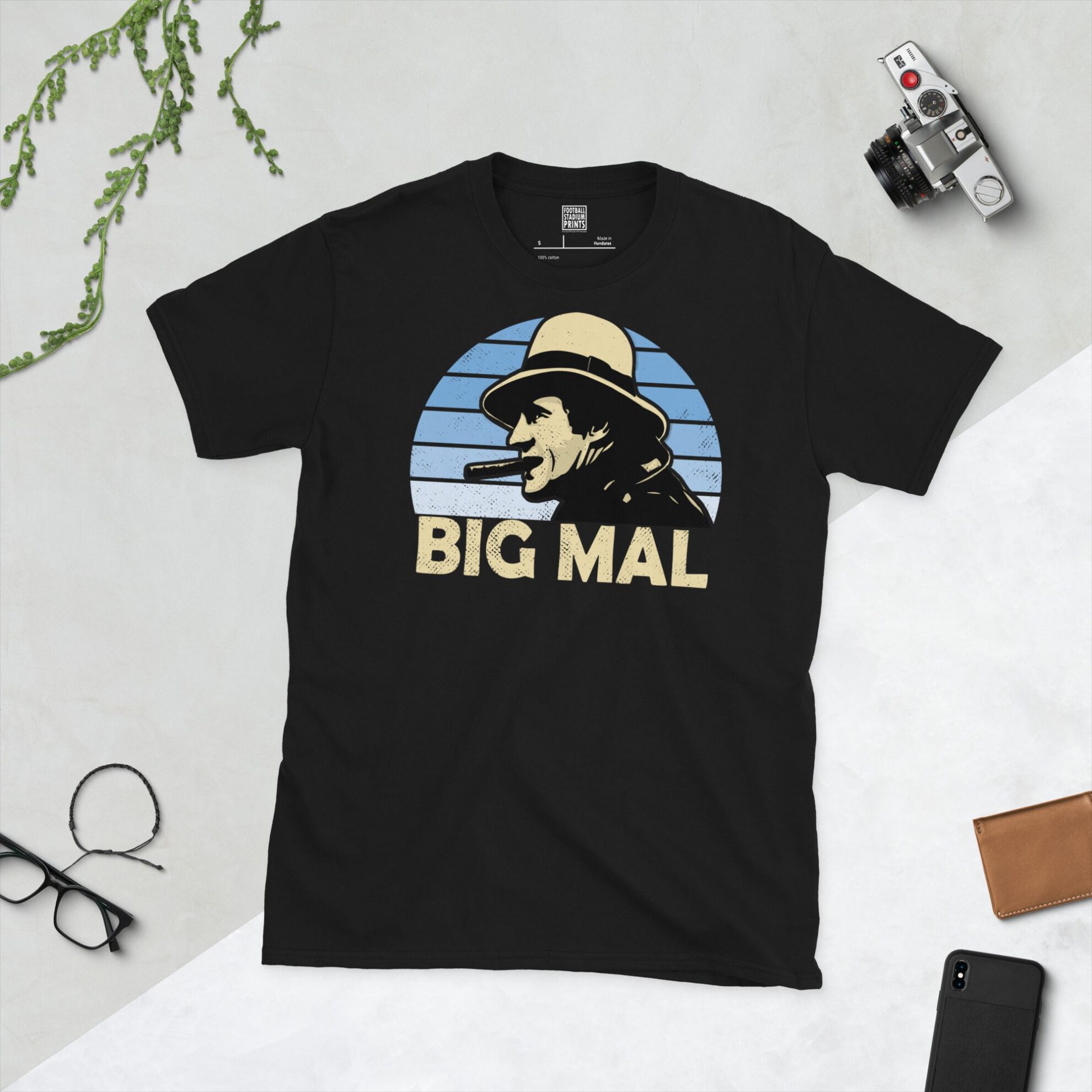 A black T-shirt featuring a stylised graphic of Manchester City legend Malcolm Allison, depicted with a fedora and cigar, against a light blue faded sunburst background. The nickname "BIG MAL" is printed in light yellow below the portrait.