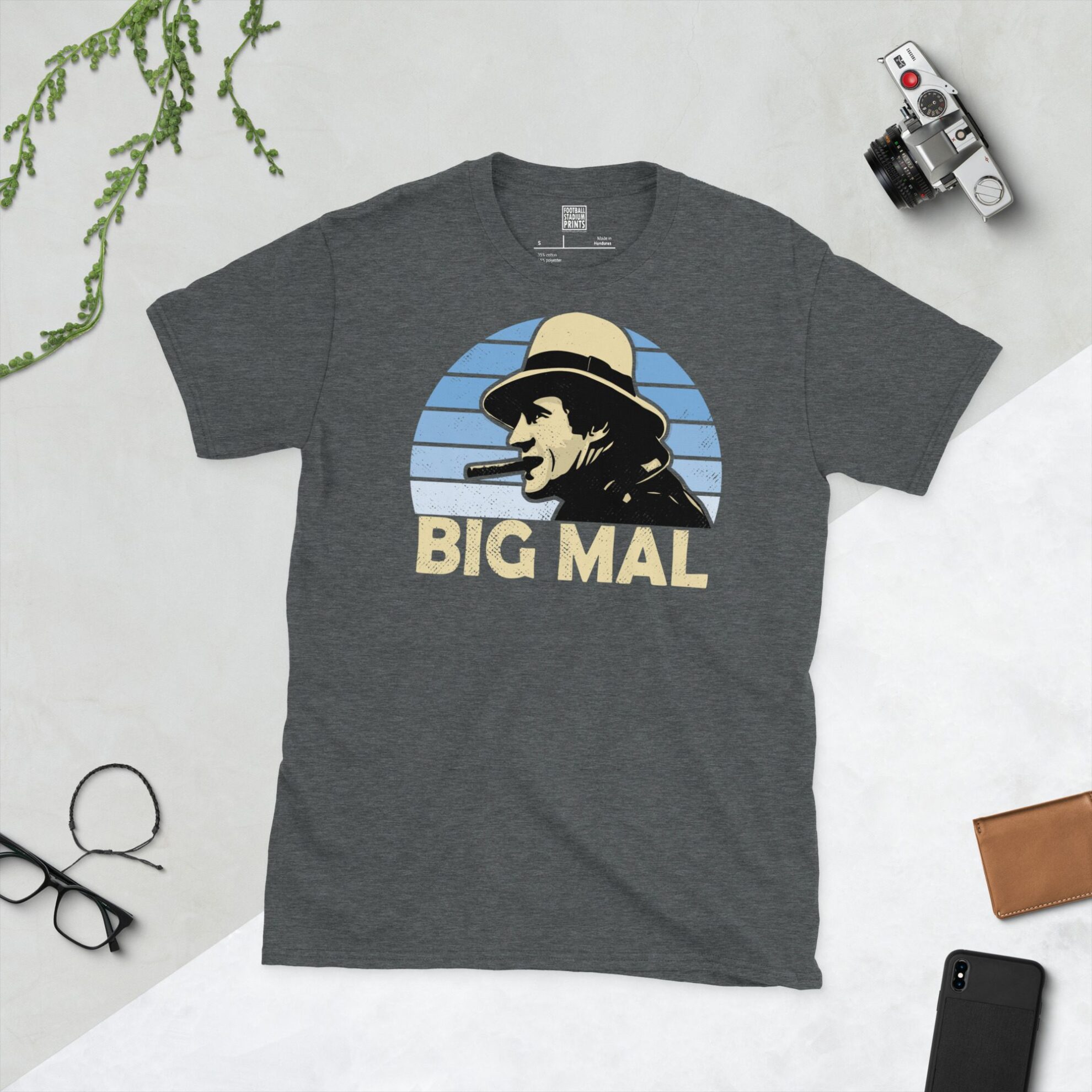 A dark heather T-shirt showing a clean, modern portrait of Malcolm Allison, the Man City icon, with "BIG MAL" printed in light yellow below the image on a light blue faded sunburst background.