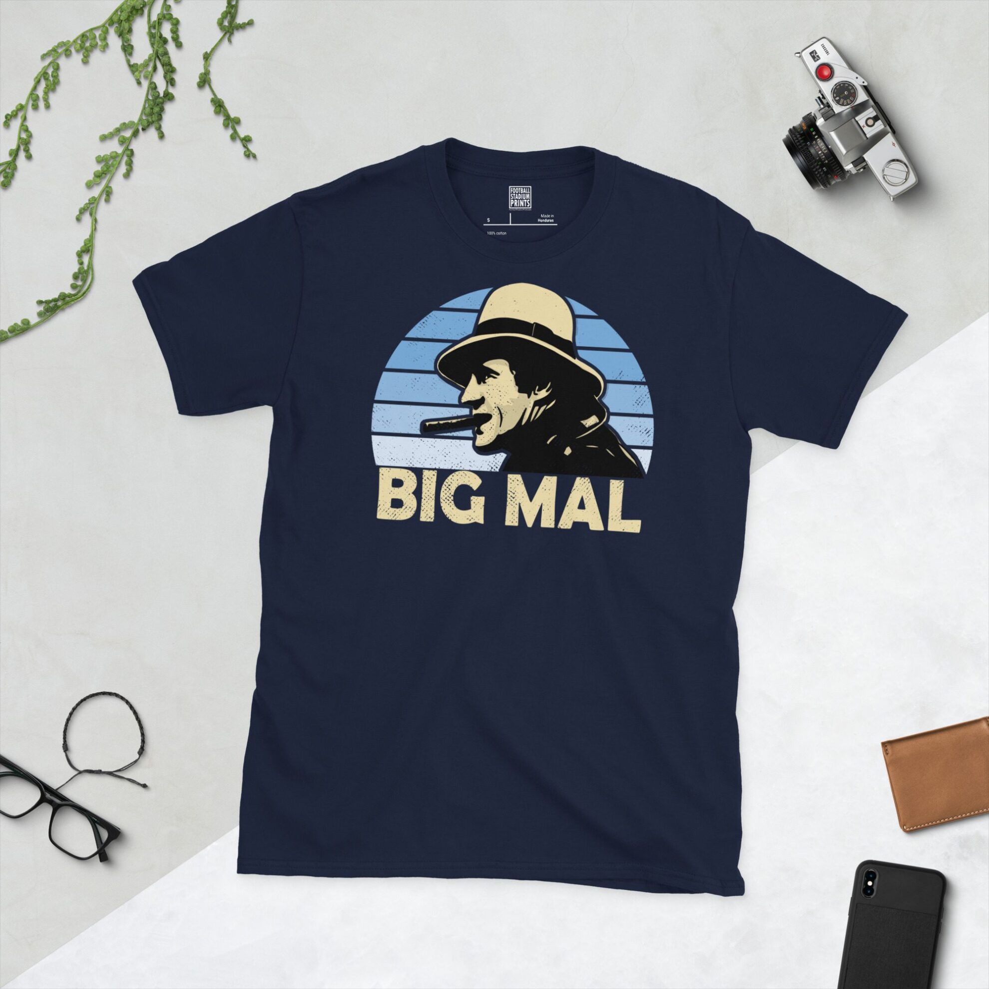 A navy T-shirt displaying an artistic illustration of Malcolm Allison, the former Man City manager. The graphic is backed by a light blue faded sunburst and the nickname "BIG MAL" appears underneath in light yellow.