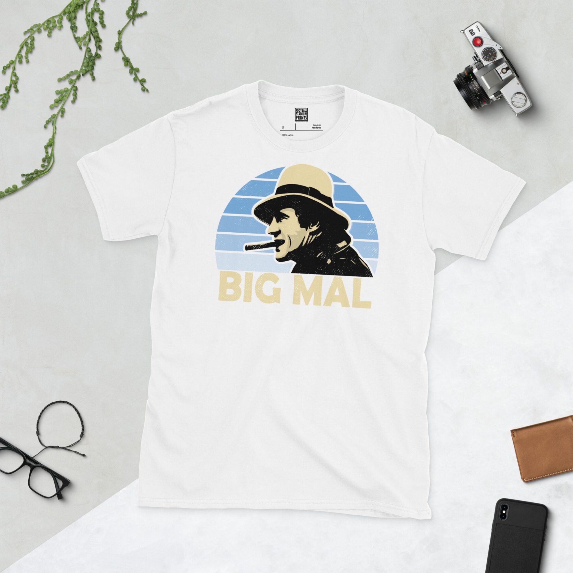 A white T-shirt with a bold portrait of football manager Malcolm Allison. The image is set against a light blue faded sunburst background with the nickname "BIG MAL" in light yellow underneath.